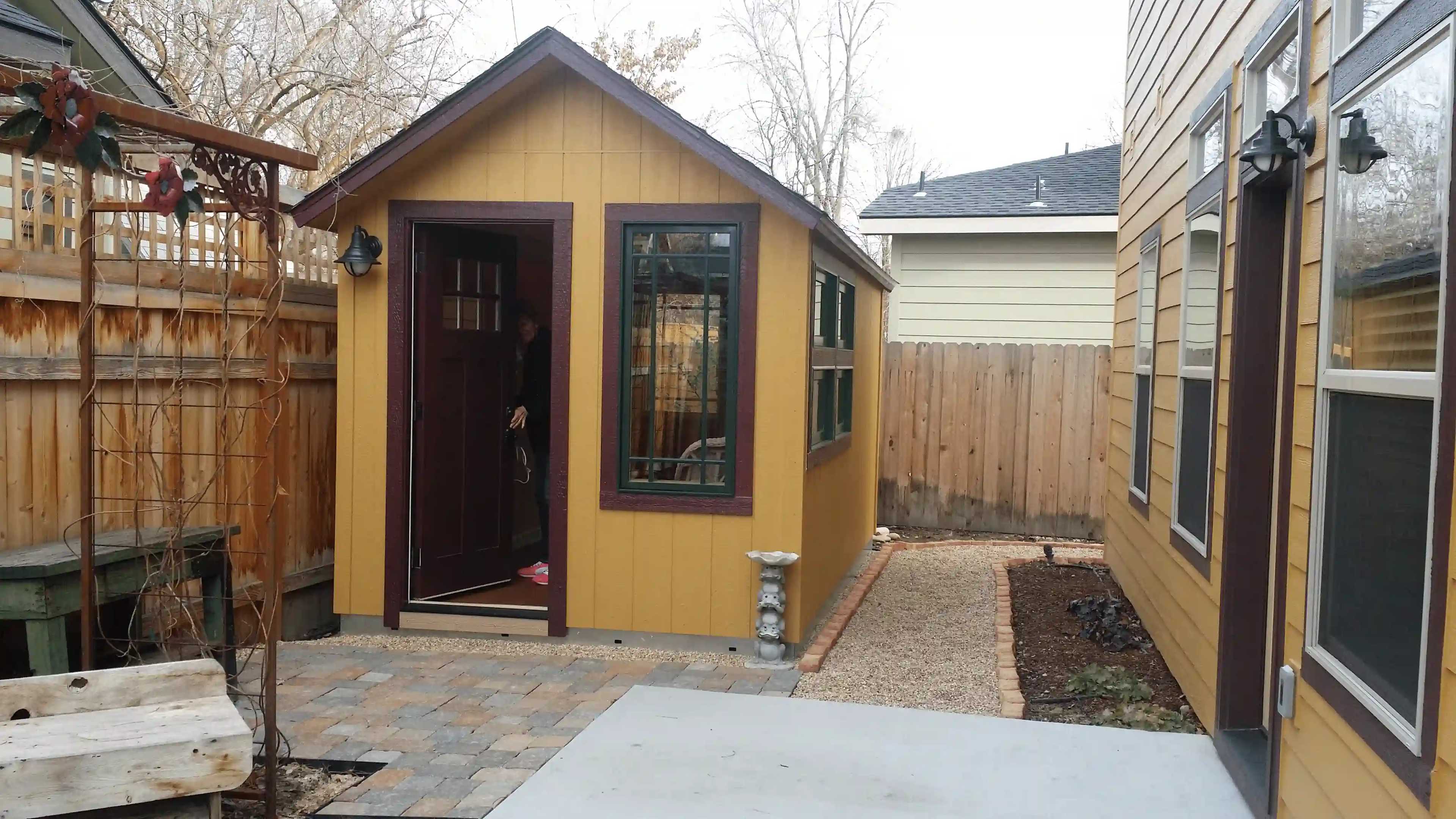 Small yellow shed with brown trim, adjacent to a house, in a fenced yard with stone and gravel pathway, surrounded by trees.