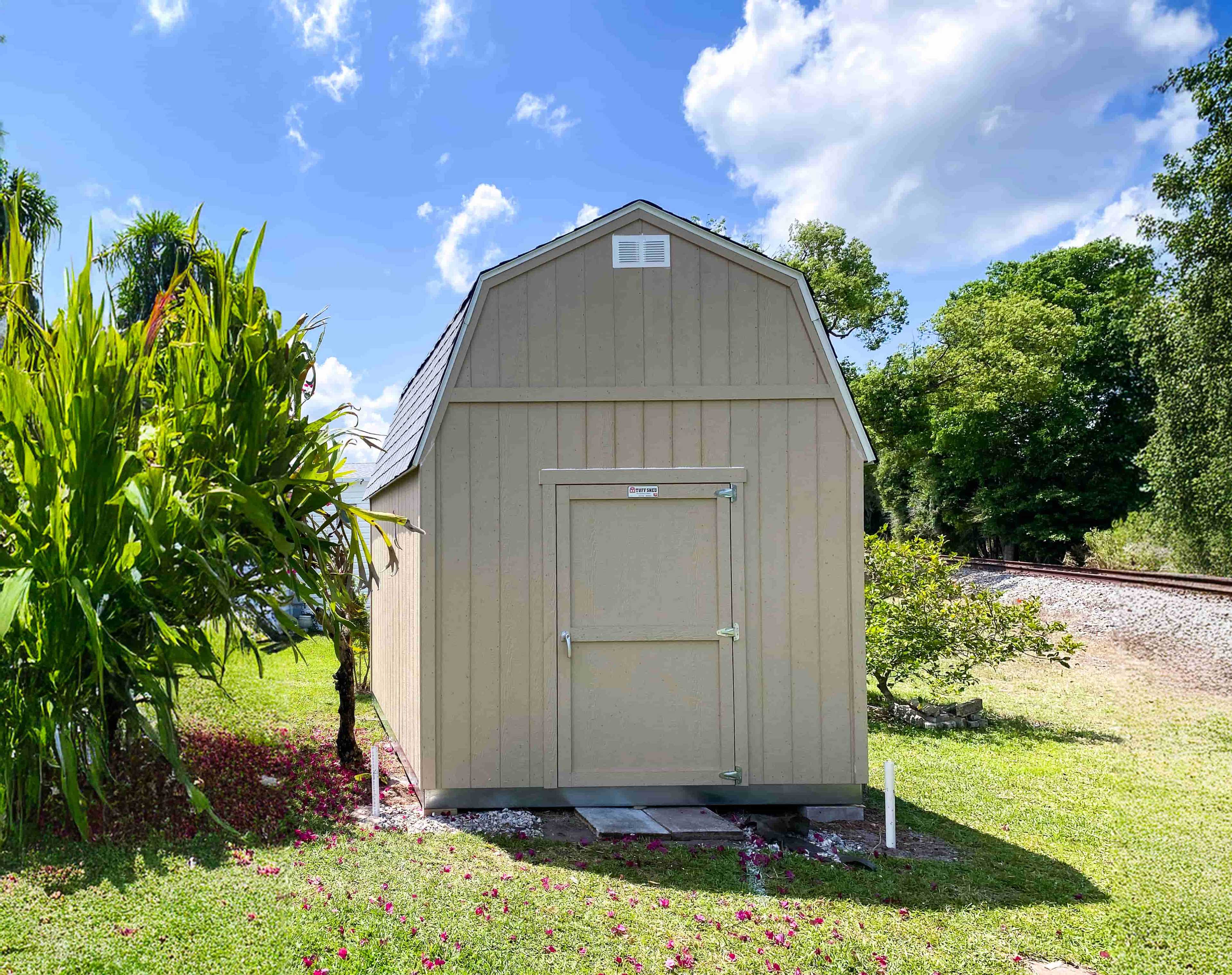 A TB700 beige shed with a gabled roof stands in a grassy area near a railroad track.