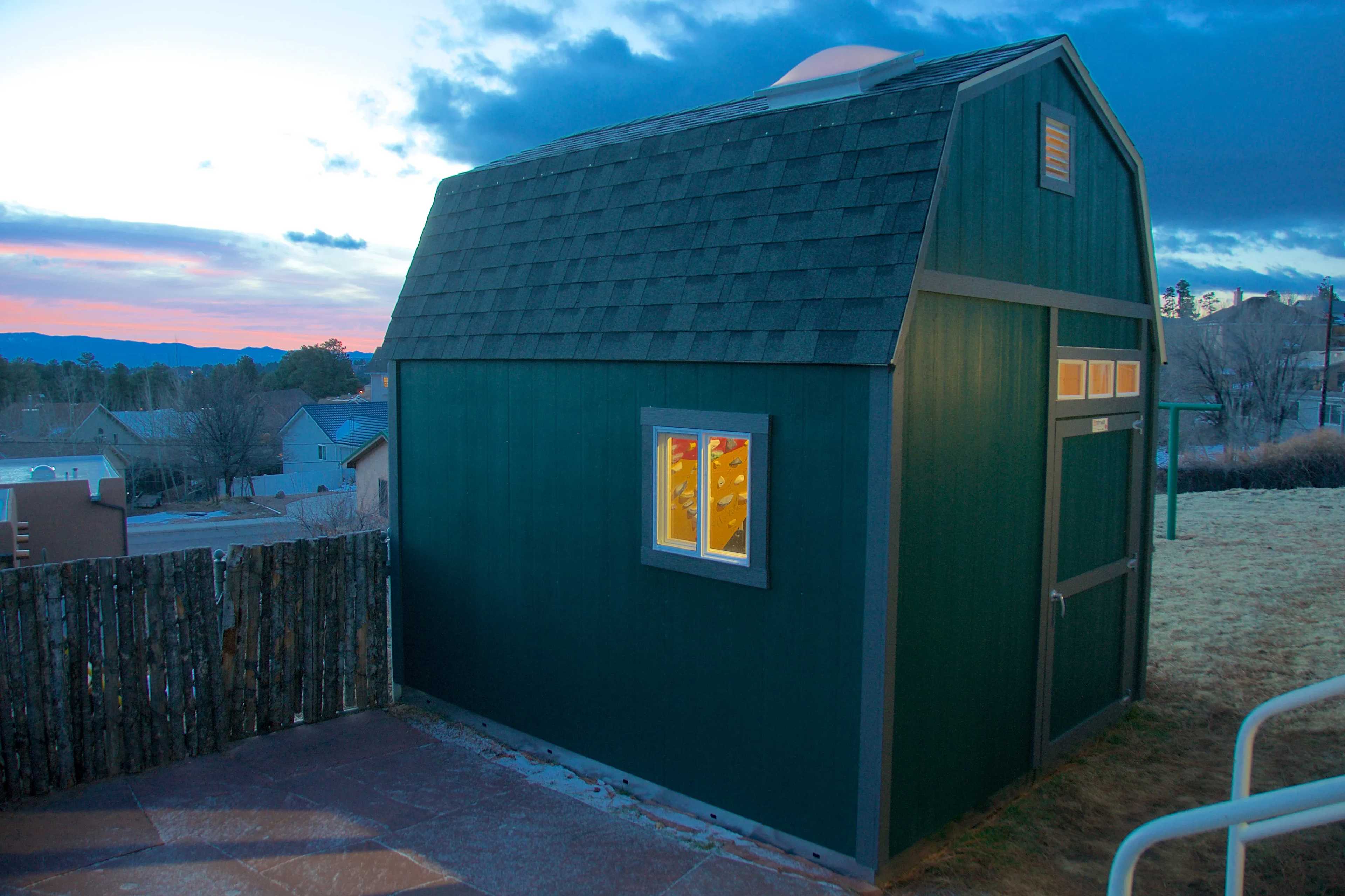A dark green shed with a shingled roof, lit from inside, stands in a yard