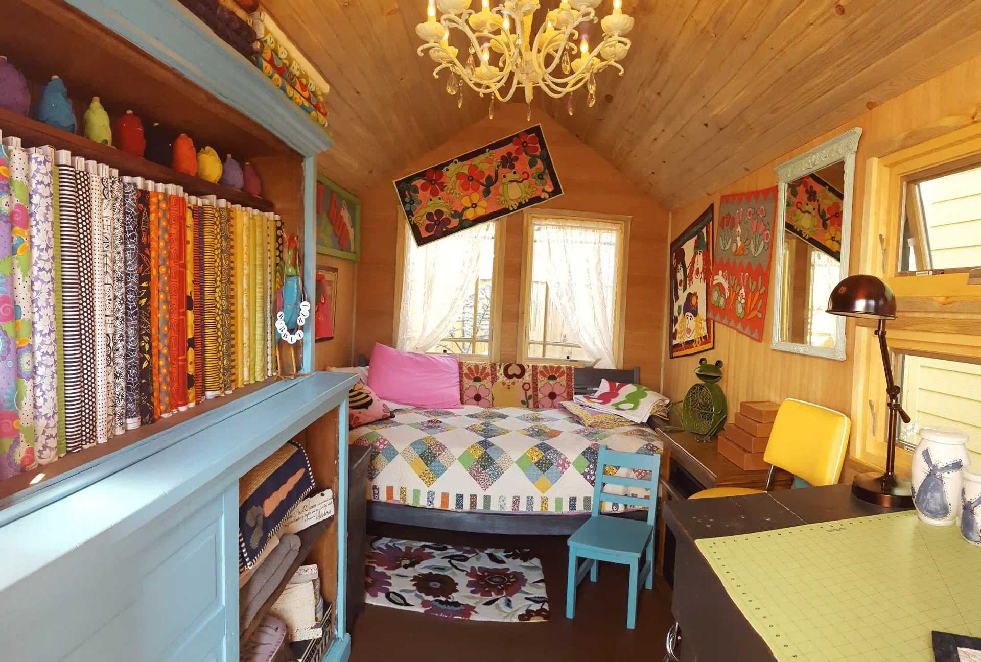 Cozy room with colorful quilts, patterned fabrics, a blue shelf, yellow-chairs, and a chandelier over a bed, creating a vibrant atmosphere.