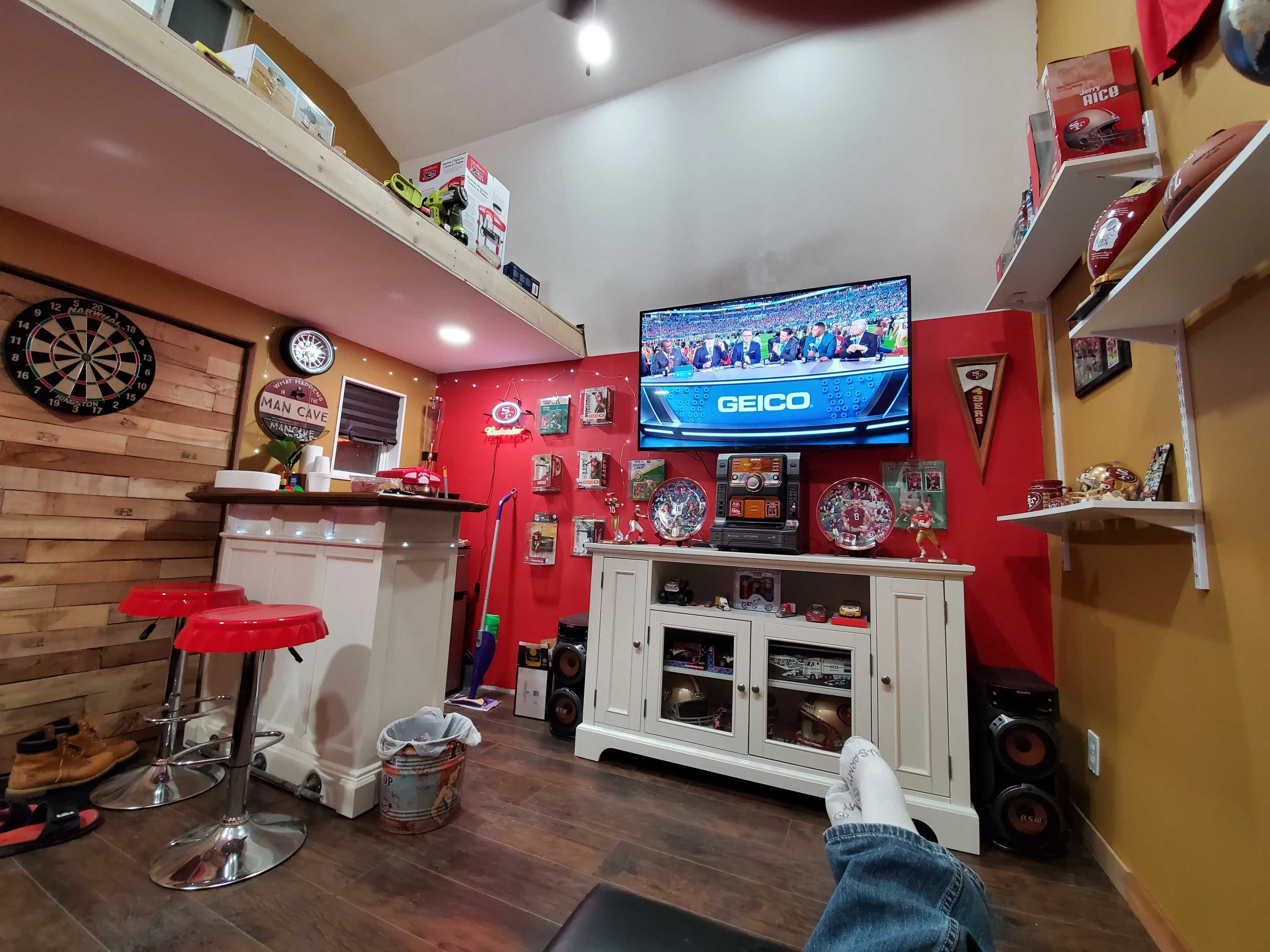 Cozy man cave with a bar, dartboard, TV showing a sports event, and various memorabilia on red and wood-paneled walls.