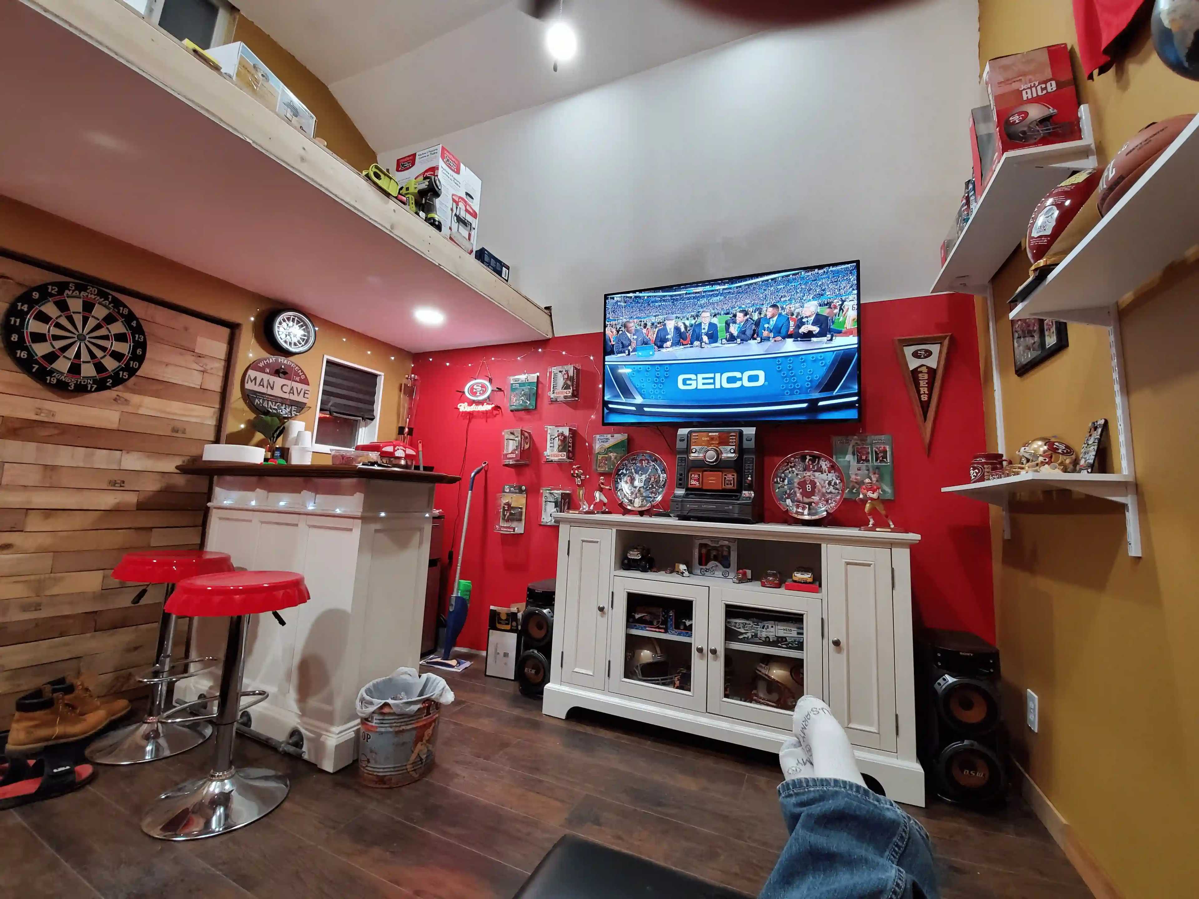Cozy man cave with bar, dartboard, red accents, TV showing a race, sports memorabilia, shelves, and a footrest in the foreground.