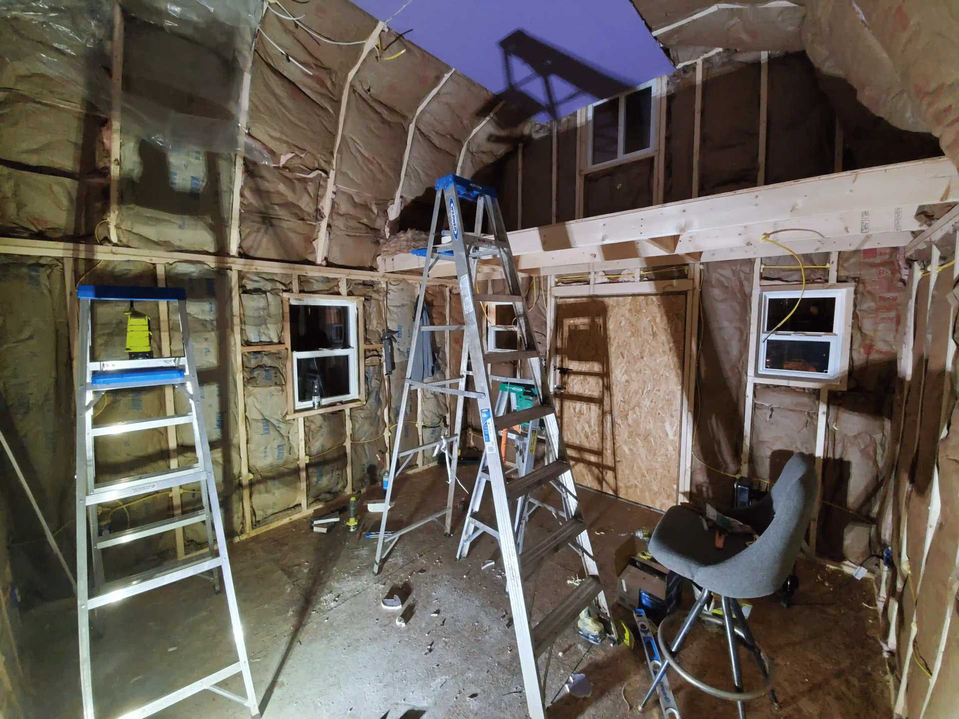 An unfinished interior space with exposed insulation and wooden framing, featuring several ladders and a chair amidst construction tools.