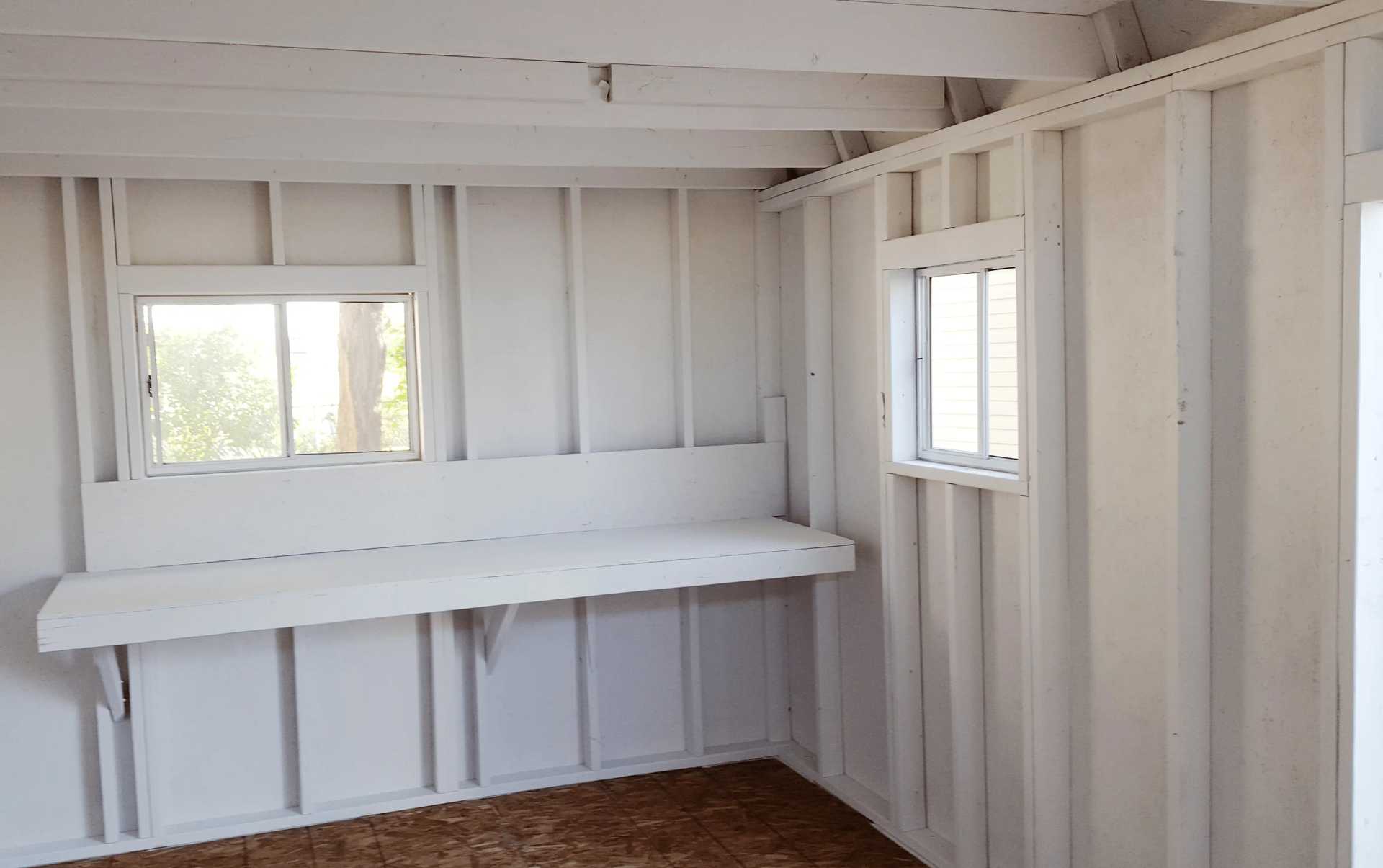 Interior of a small, unfinished room with white wooden walls, two windows, and a narrow shelf installed beneath one window.