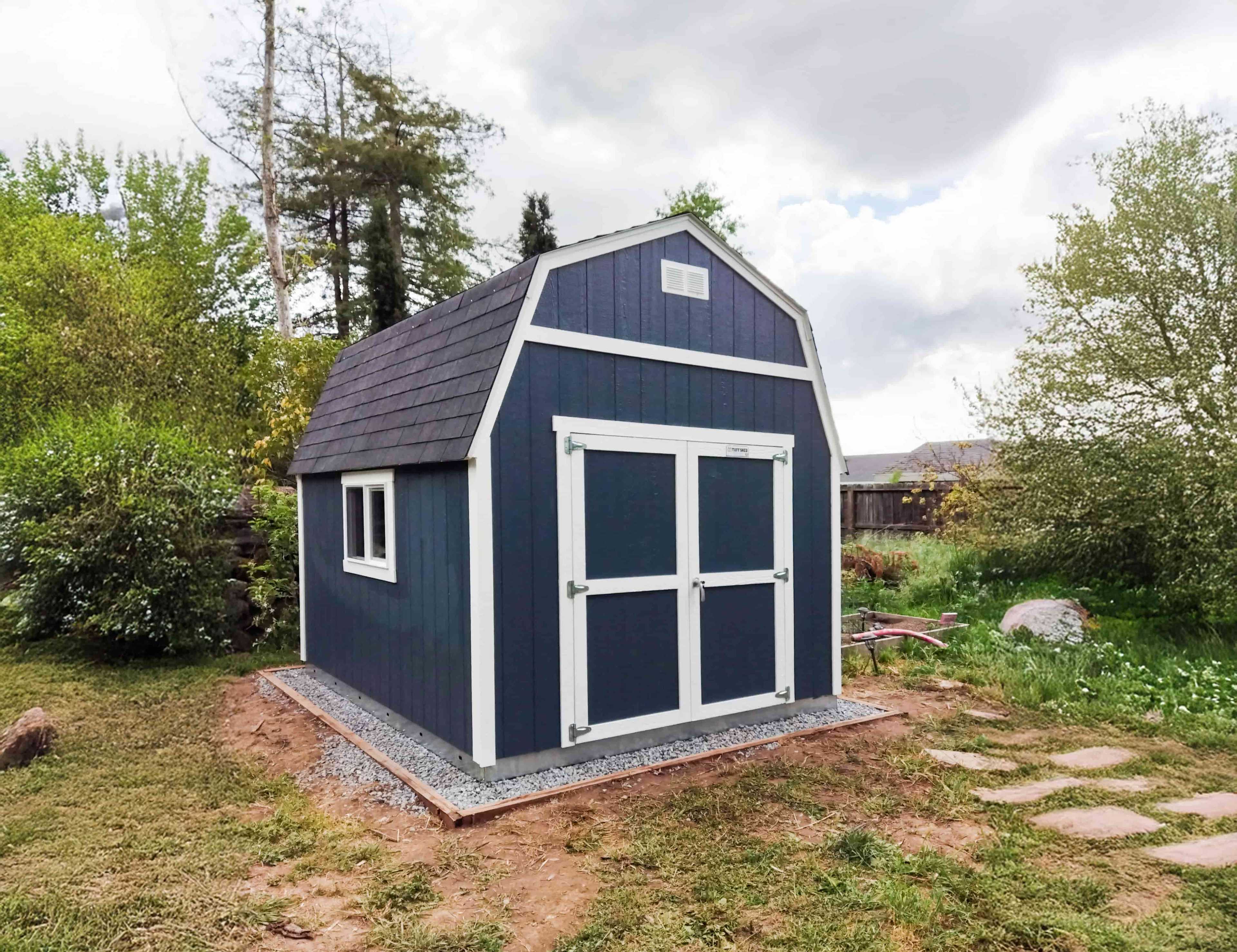A blue TB600shed with white trim and a gabled roof stands on a gravel base in a grassy backyard