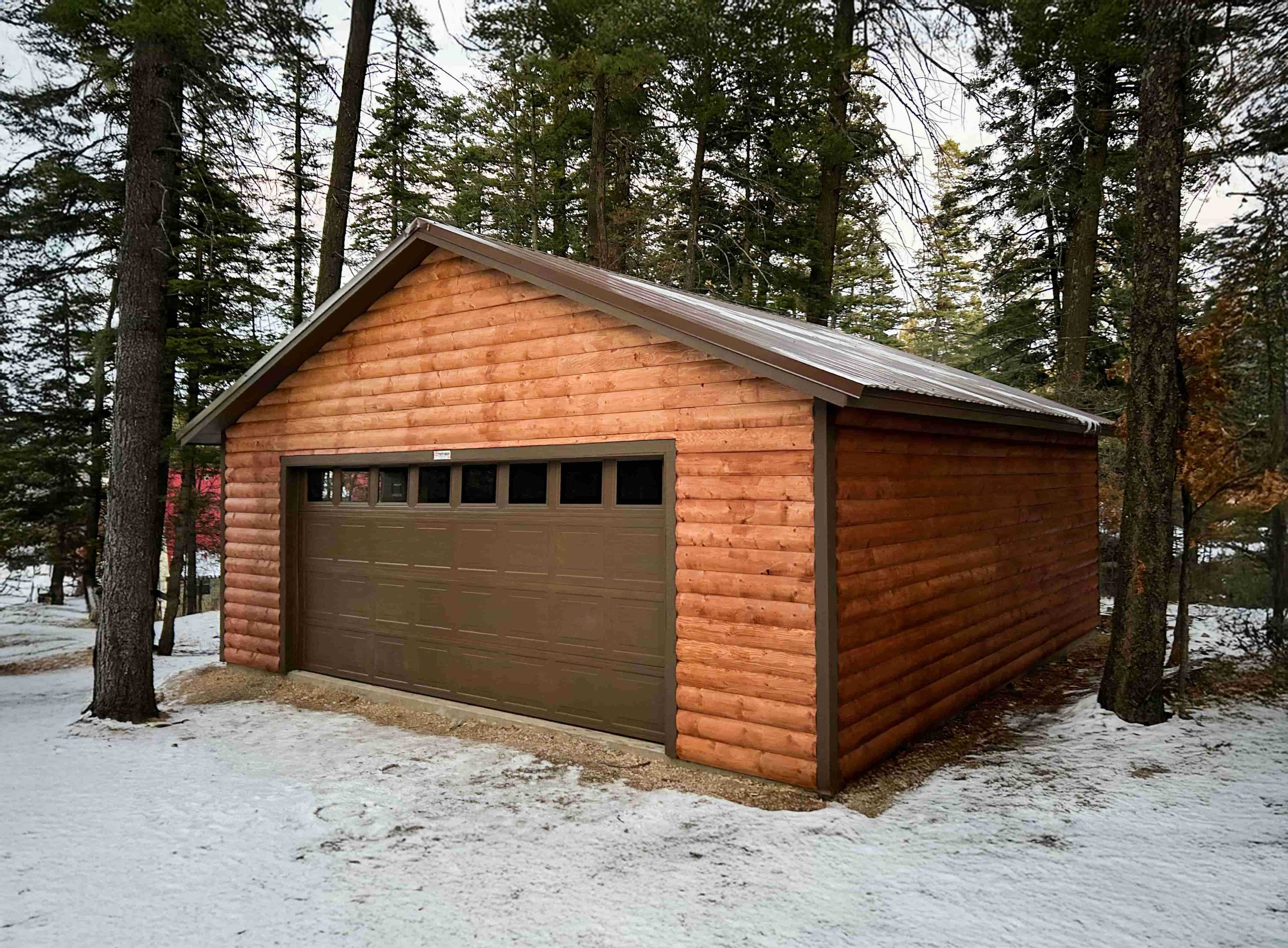 A wooden Sundance Ranch Garage with a brown door sits in a snowy forested area, surrounded by tall trees
