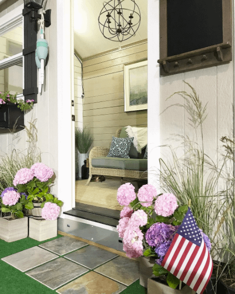 Cozy porch with wicker seating, pink hydrangeas, and an American flag. Hanging light fixture and potted plants add charm.