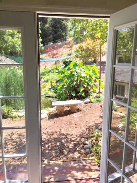 Open double doors lead to a sunny garden with a stone bench, lush greenery, and a small hill in the background.