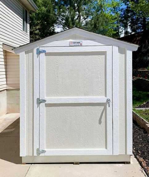 White SR600 outdoor storage shed with a pitched roof, located on a concrete patio next to a house