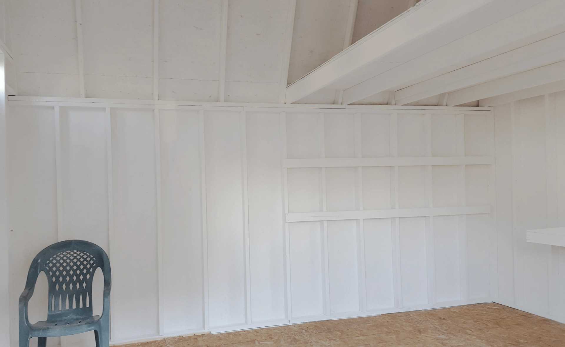 Empty white room with wooden panel walls, a plastic chair on the left, and two simple shelves on one wall. Ceiling beams are visible.