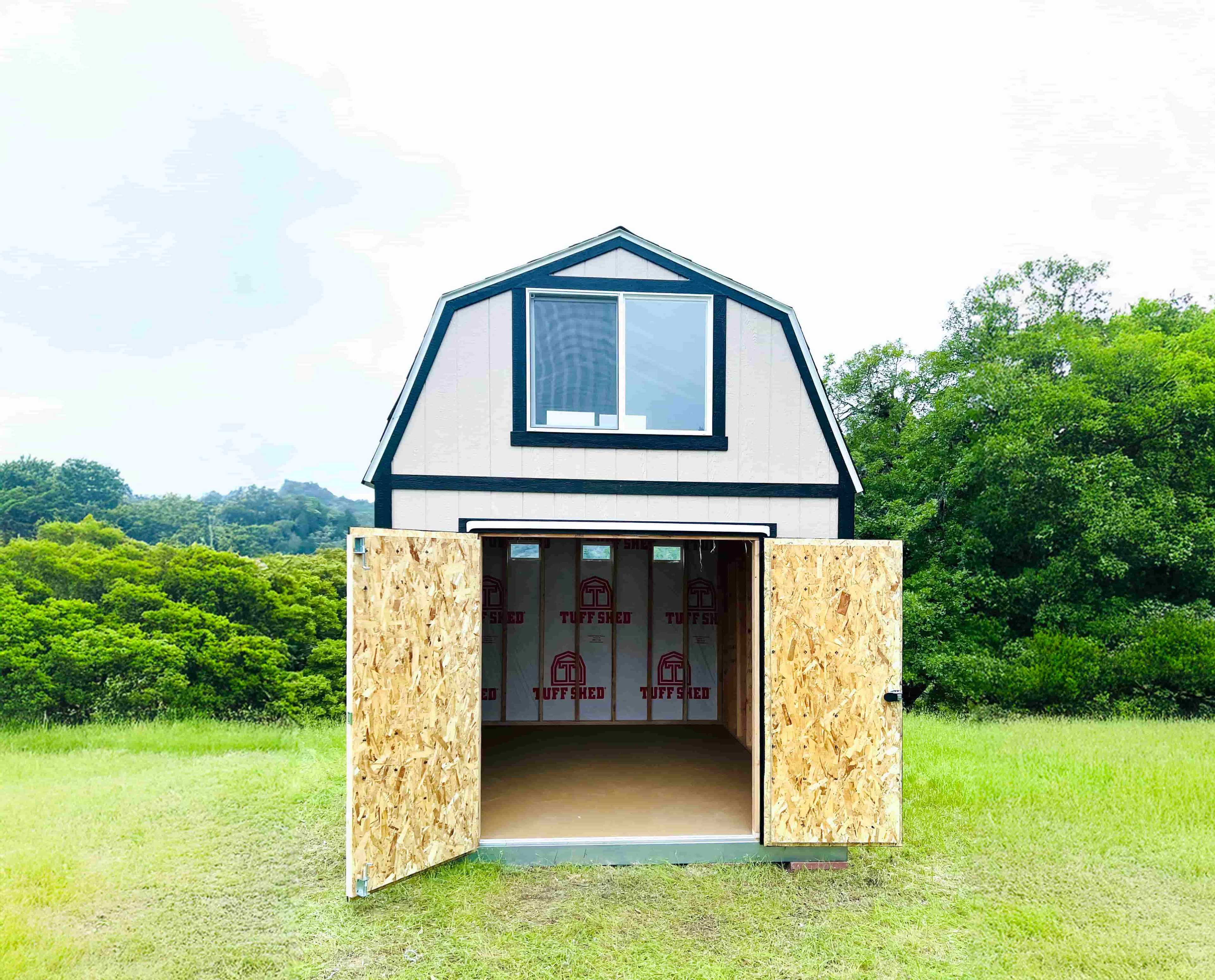 TB800 barn-style shed with open doors, revealing an unfinished interior. Set on a grassy field with trees and hills in the background.