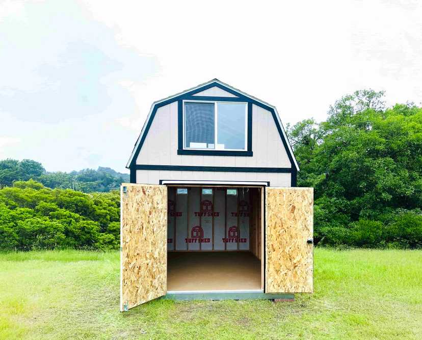 TB800 barn-style shed with open doors, revealing an unfinished interior. Set on a grassy field with trees and hills in the background.