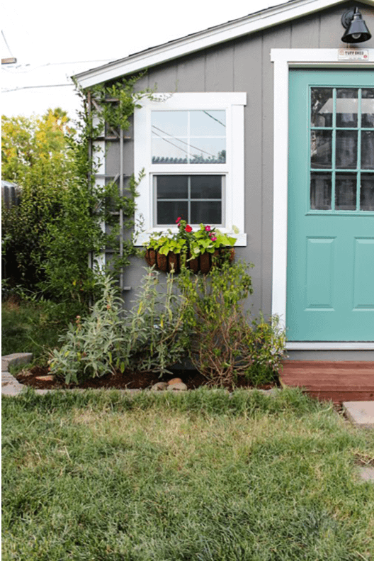 SR600 Exterior She Shed Makeover 2 SR600 shed with a teal door, white-trimmed window, potted plants, and lush greenery in a grassy yard.