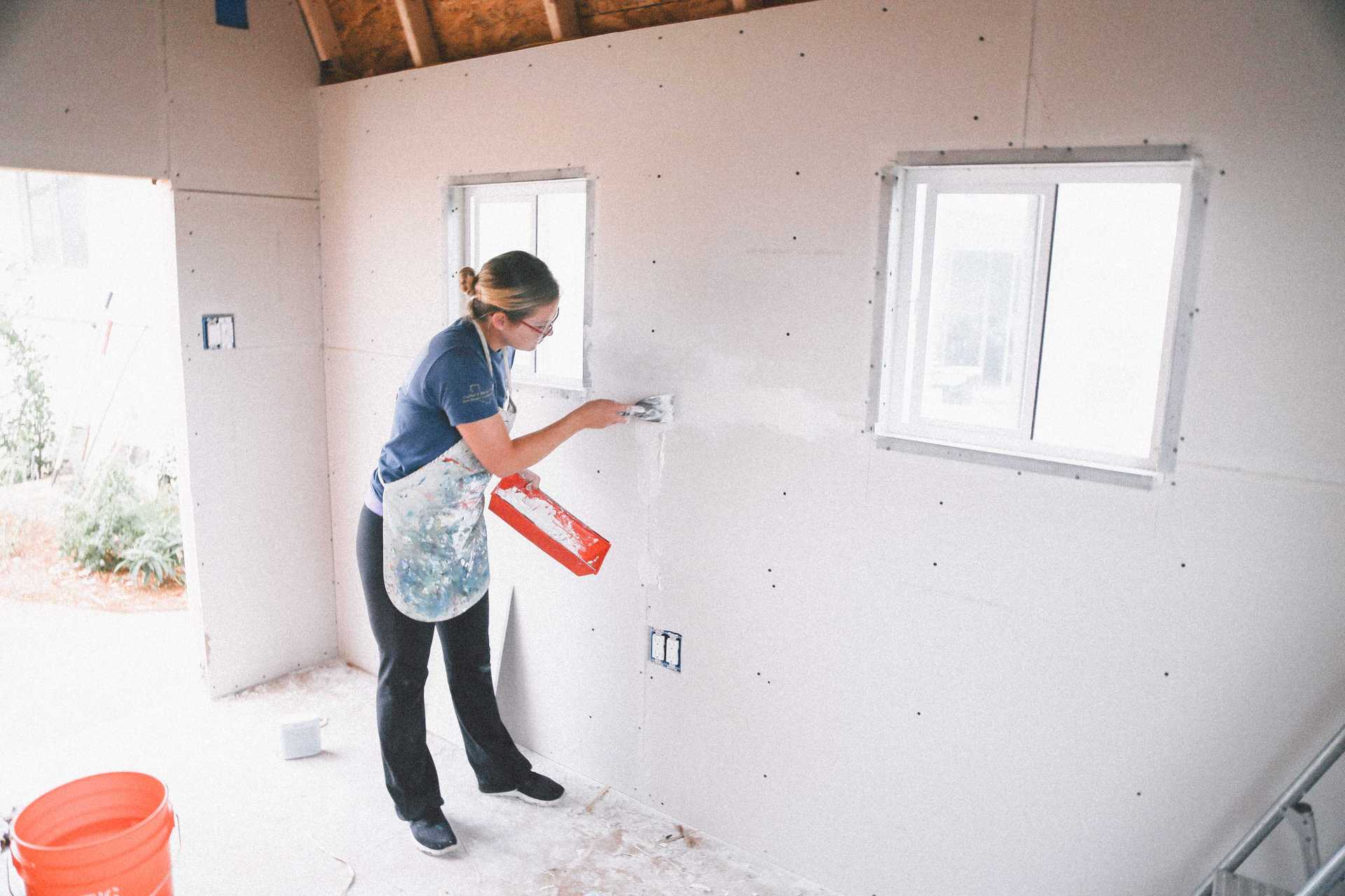 Michaela finishing out the interior of her new TB-700 A woman applies joint compound to drywall seams in a room under construction, wearing a blue shirt and apron, with windows in the background.