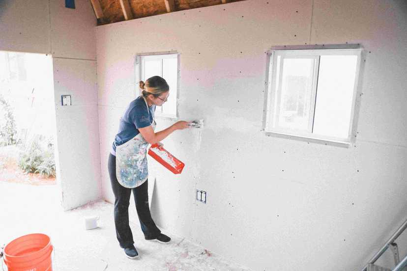 A woman patching the drywall in wearing a blue shirt and apron, with a window in the background.
