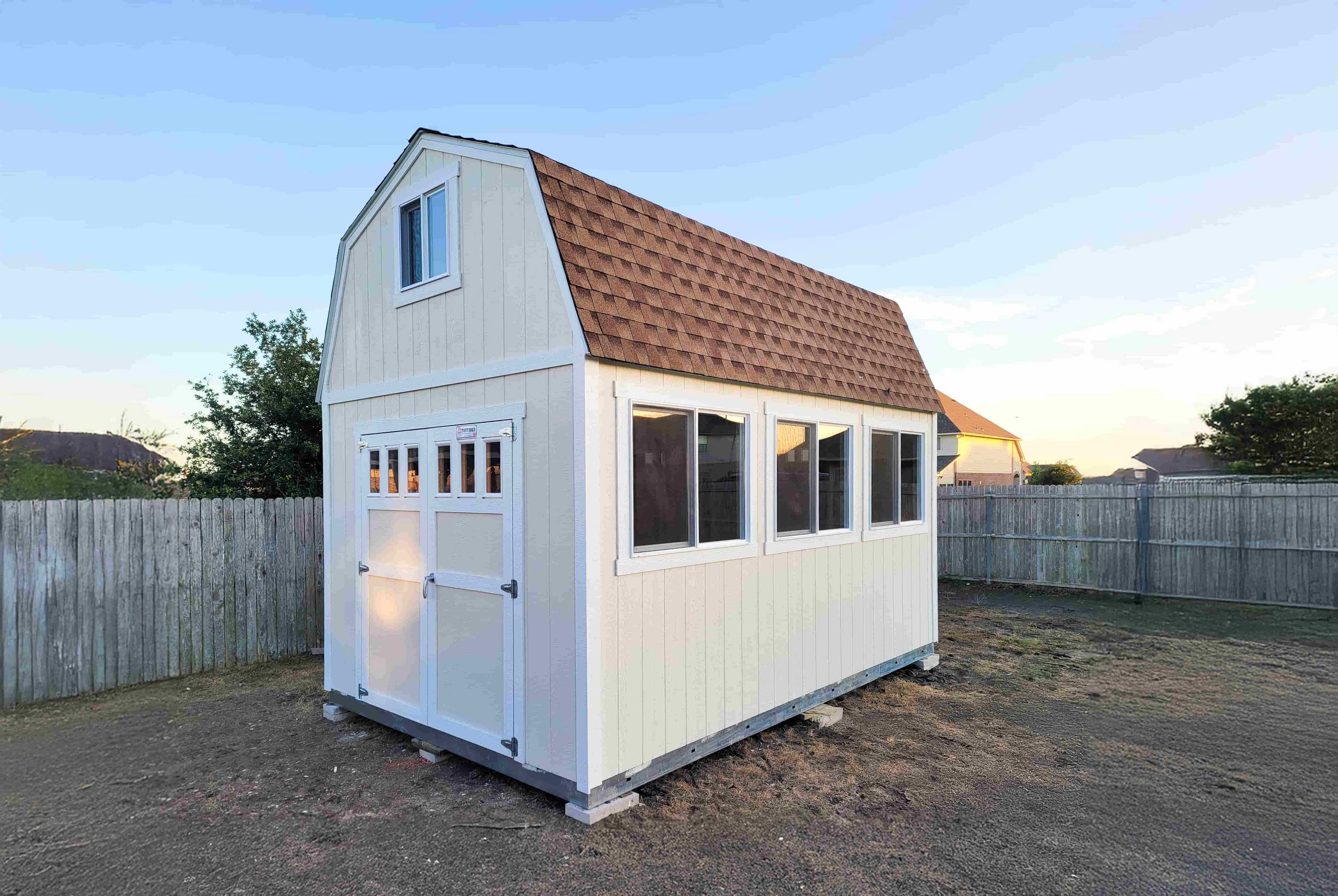 A beige shed with a brown shingled roof and multiple windows stands on a dirt patch, surrounded by a wooden fence.