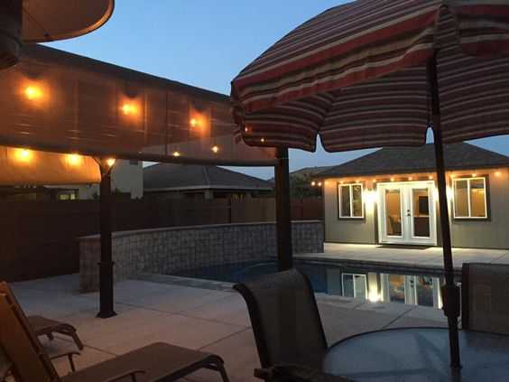 Poolside patio at dusk with string lights, striped umbrella, and lounge chairs. A Tuff Shed light up in the background