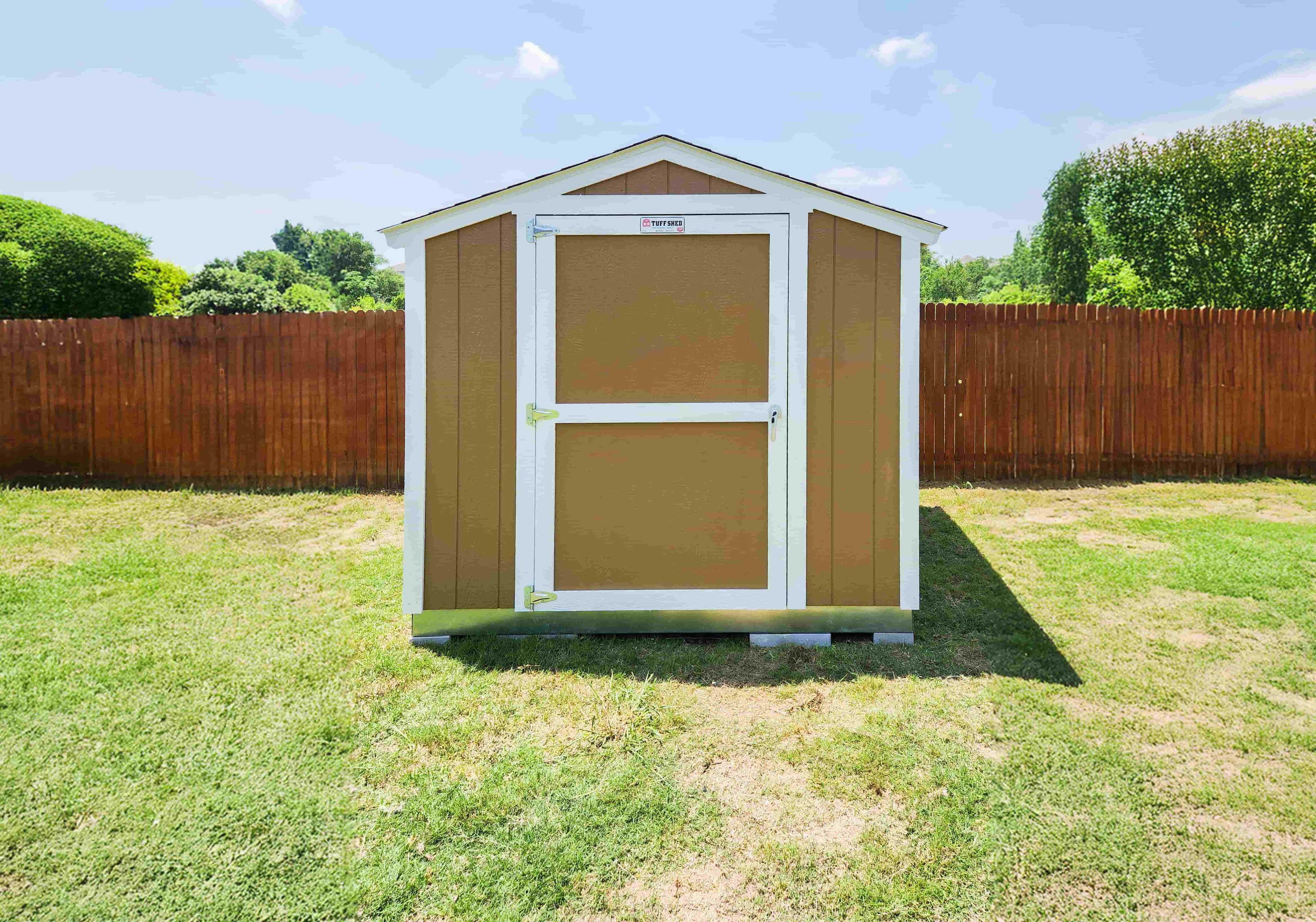 A SR600 brown and white shed with a closed door, set in a grassy yard with a wooden fence and trees in the background under a clear sky.