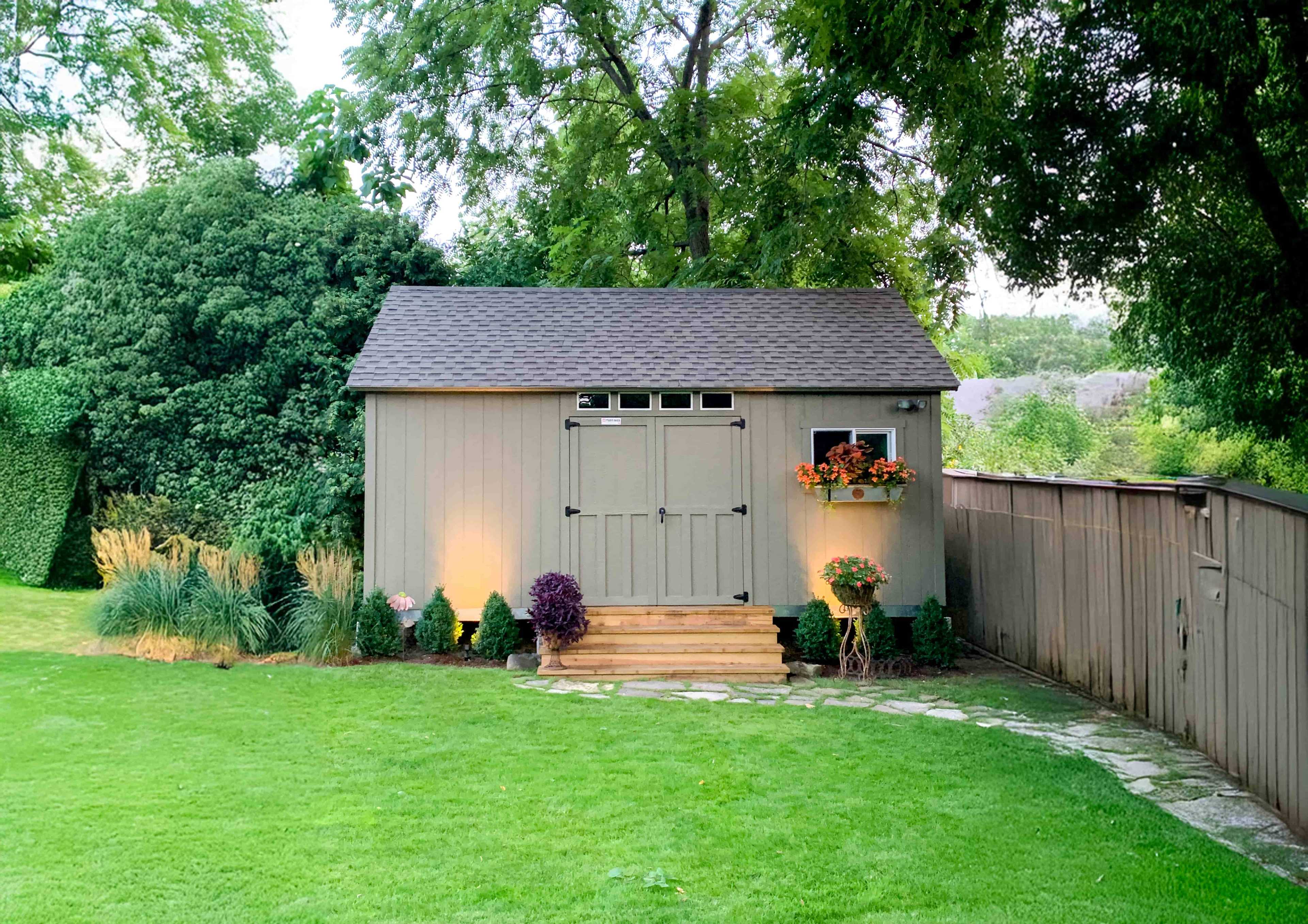 ATR800 garden shed with gray siding, lit by ground lights, surrounded by greenery, with a flower box under a window and a wooden step entrance.