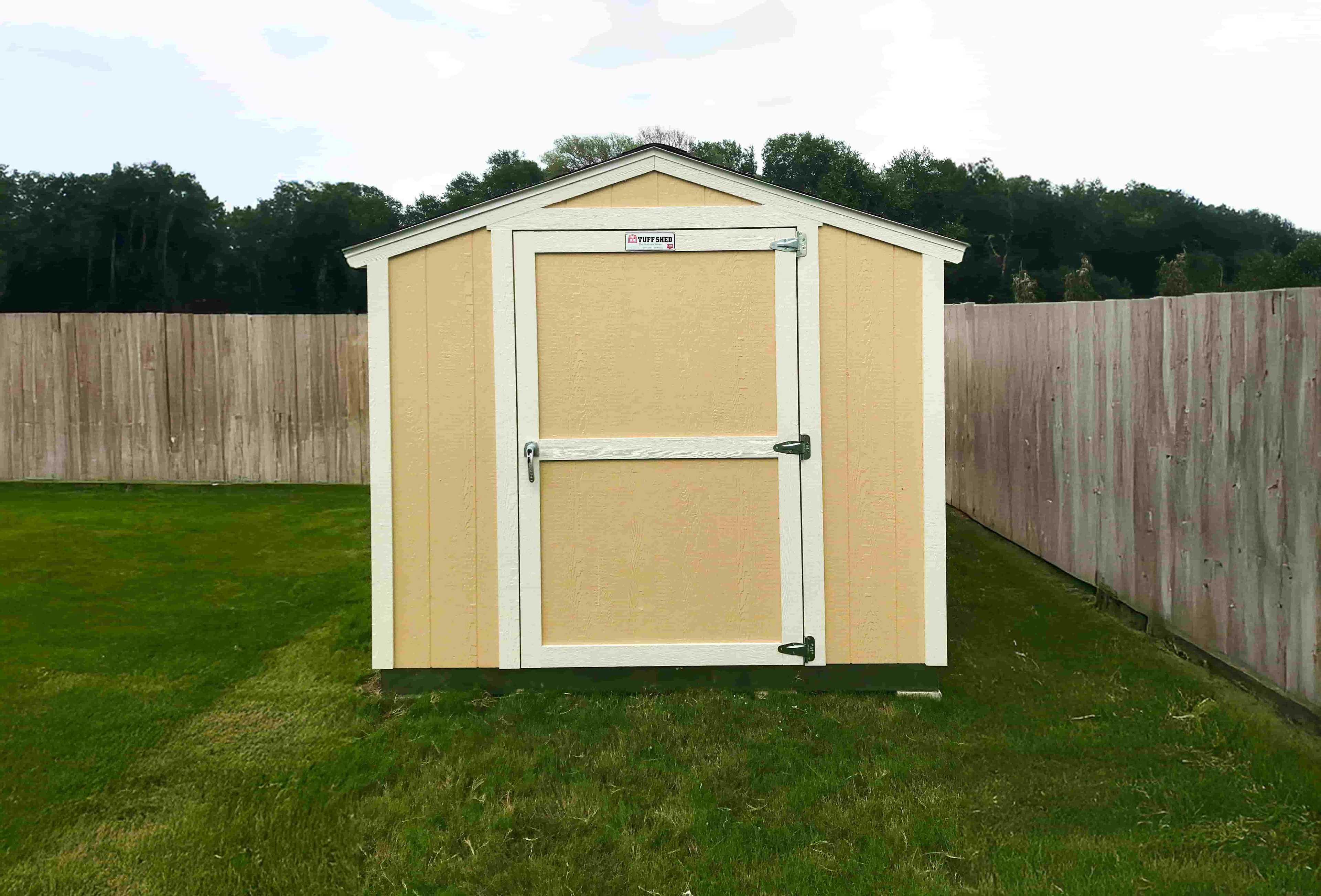 A beige and white SR600 storage shed with a single door stands on a grassy lawn, enclosed by a wooden fence and trees in the background.