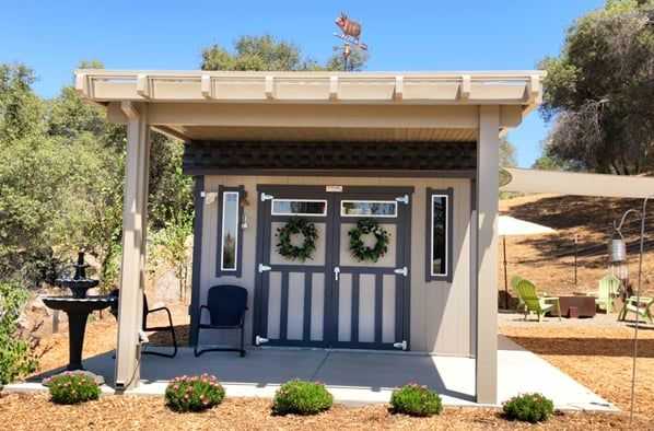 Small shed with double doors adorned with wreaths, surrounded by trees and garden chairs, under a clear blue sky.