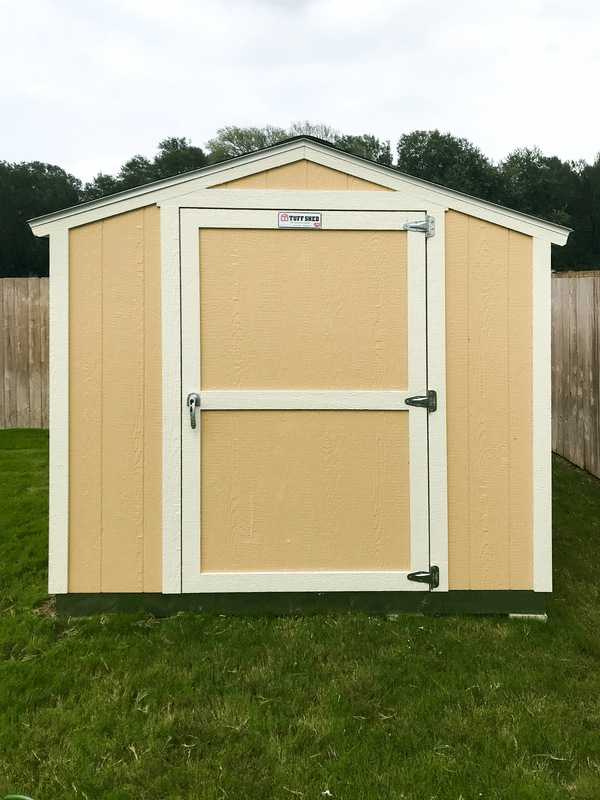 A beige and white SR600 storage shed with a single door stands on a grassy lawn, enclosed by a wooden fence and trees in the background.