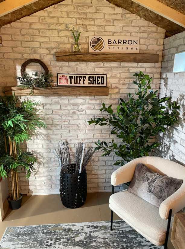 Cozy corner with a white brick wall, green plants, a beige chair, and decorative signs for Barron Designs and Tuff Shed.