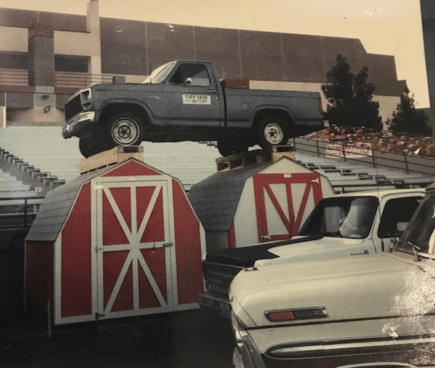 Tuff Sheds Tough Enough for a Truck A truck is parked atop two red and white sheds in a stadium setting, with other vehicles visible below.