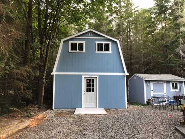A TB800 barn-style shed with white trim stands in a forested area, next to a smaller shed and a patio set on gravel.