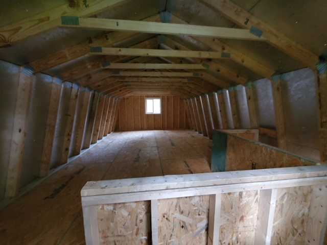 Interior of an unfinished attic with exposed wooden beams, plywood floor, and a small window at the far end, under construction.