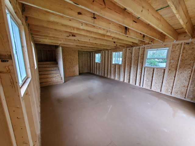 Unfinished wooden interior of a house with exposed beams, plywood walls, windows, and a staircase on the left.