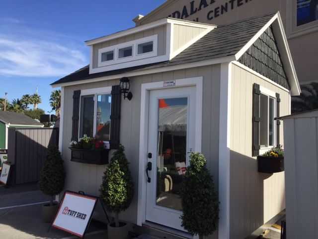 Tuff Shed with a white door, black shutters, and flower boxes. It has a gabled roof and is displayed outdoors with signage.