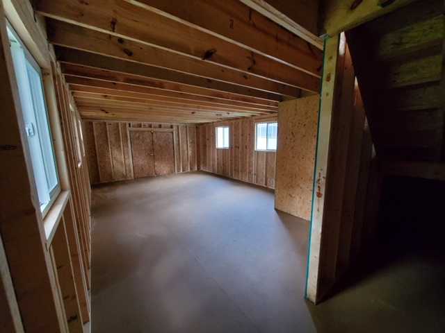 Unfinished basement with exposed wooden beams and walls, two small windows, and a staircase leading up on the right.