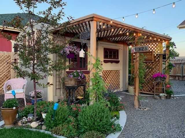 Charming garden shed with wooden pergola, hanging plants, and string lights. Surrounded by lush greenery and a gravel path.