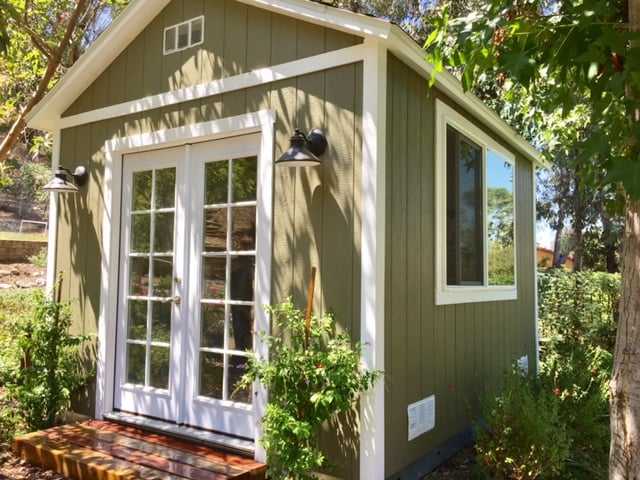 A TR800 shed with French doors, surrounded by trees and plants, in a sunny garden setting