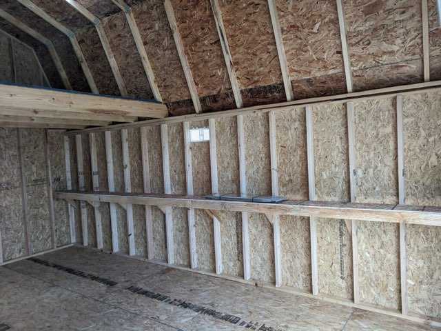 Interior of an unfinished TB700 wooden shed with exposed studs, a small window, and a loft area.