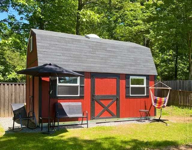 Red barn-style shed with a black roof in a backyard. Two chairs, a hammock, and an umbrella are nearby. Lush green trees in the background.