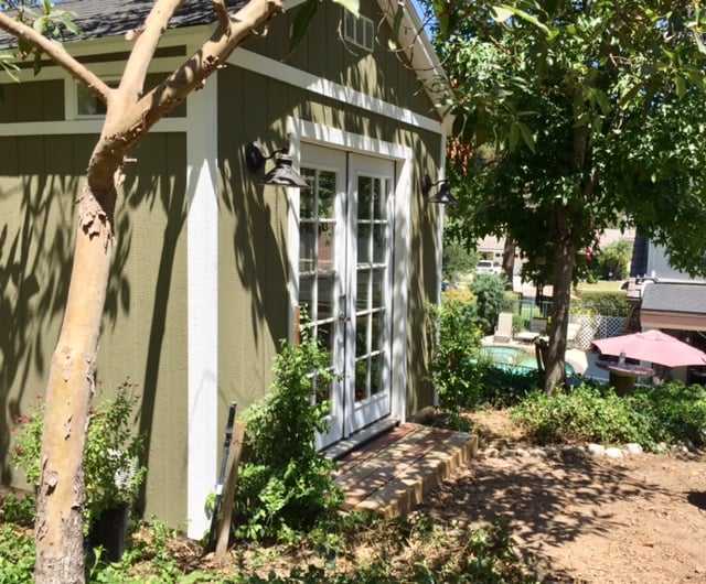Green TR800 garden shed with white French doors, surrounded by trees and plants, under a sunny sky