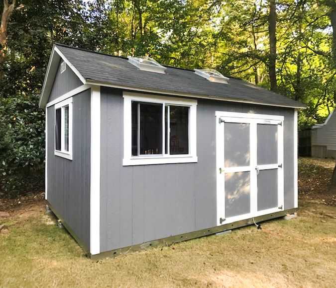 A gray TR800 garden shed with white trim, two windows, and double doors, set on grass with trees in the background.