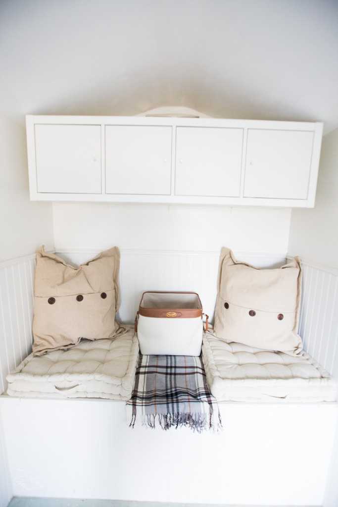 Cozy nook with beige cushions, white storage cabinets above, and a plaid blanket draped over a bench with a brown bag on it.