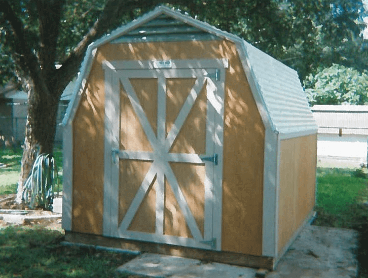 40th Anniversary Tuff Shed Building Example 2 A wooden shed with a metal roof and double doors, set on a concrete slab, surrounded by grass and a large tree.