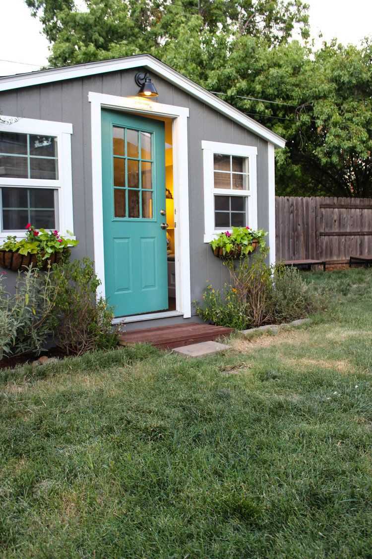 Small gray shed with a teal door, white trim, and flower boxes. Warm light glows inside. Surrounded by grass and a wooden fence.