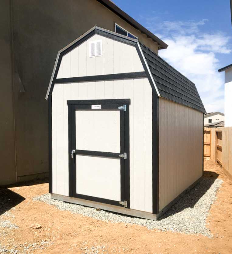 A beige shed with a black roof and trim stands on a gravel base between two buildings under a clear blue sky.