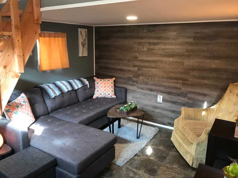 Cozy TB600 shed with a gray sectional sofa, patterned cushions, wicker chair, wooden wall, and small coffee table on a rug.