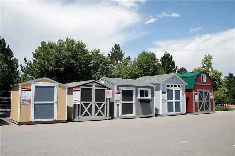 A row of various outdoor storage Tuff Shed buildings displayed on a paved area, surrounded by trees under a partly cloudy sky.