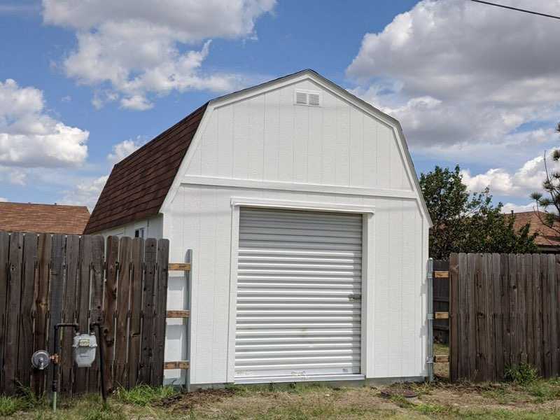 A white barn-style TB700 shed with a brown roof and a closed metal roll-up door, surrounded by a wooden fence under