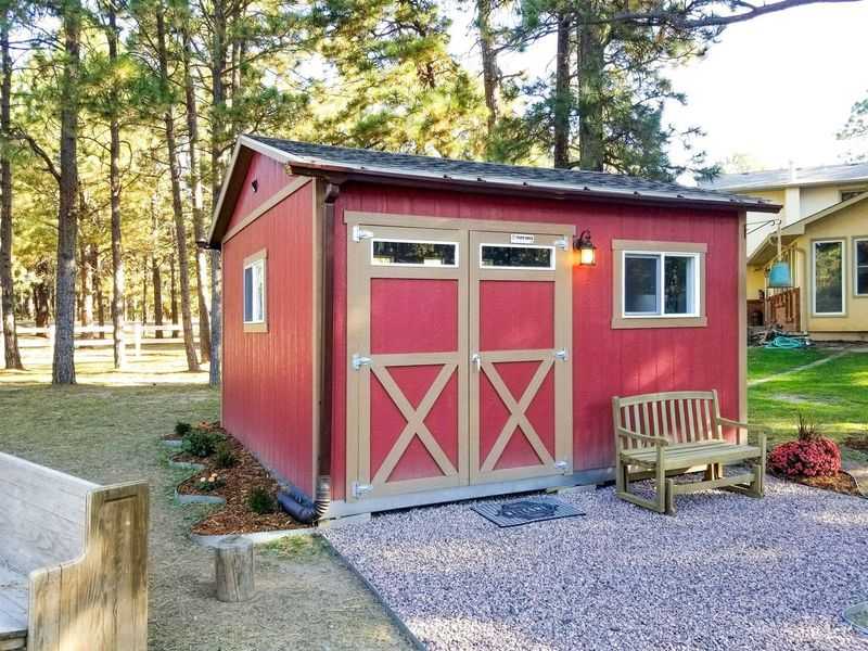 A red TR800 shed with double doors, surrounded by trees, a bench, and gravel path, next to a house in a forested area.