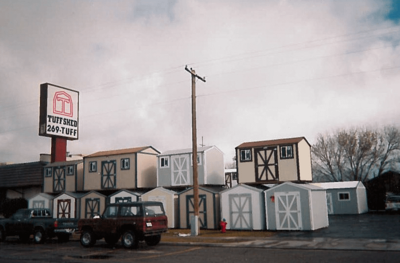 An early years display of Tuff Shed Buildings Outdoor display of various storage sheds and barns under a cloudy sky, with a "Tuff Shed" sign and parked vehicles nearby.