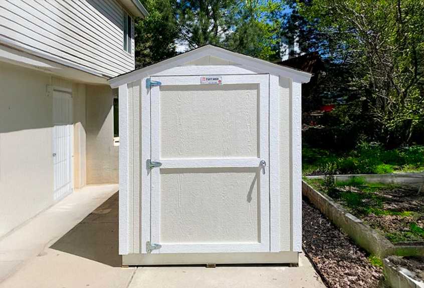 White SR600 outdoor storage shed with a pitched roof, located on a concrete patio next to a house
