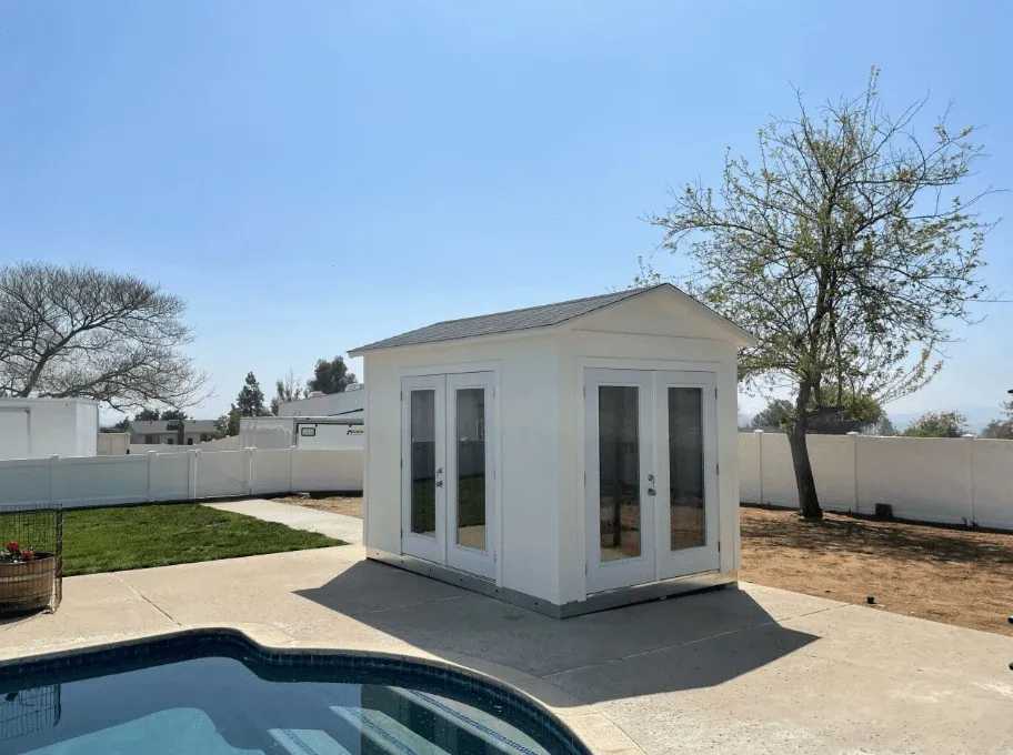 Small white shed with glass doors beside a pool, surrounded by a concrete patio, a grassy area, and trees under a clear blue sky.